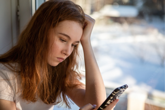 Teen girl looking worried while sitting by a window and looking at her smartphone, illustrating distress related to online interactions.